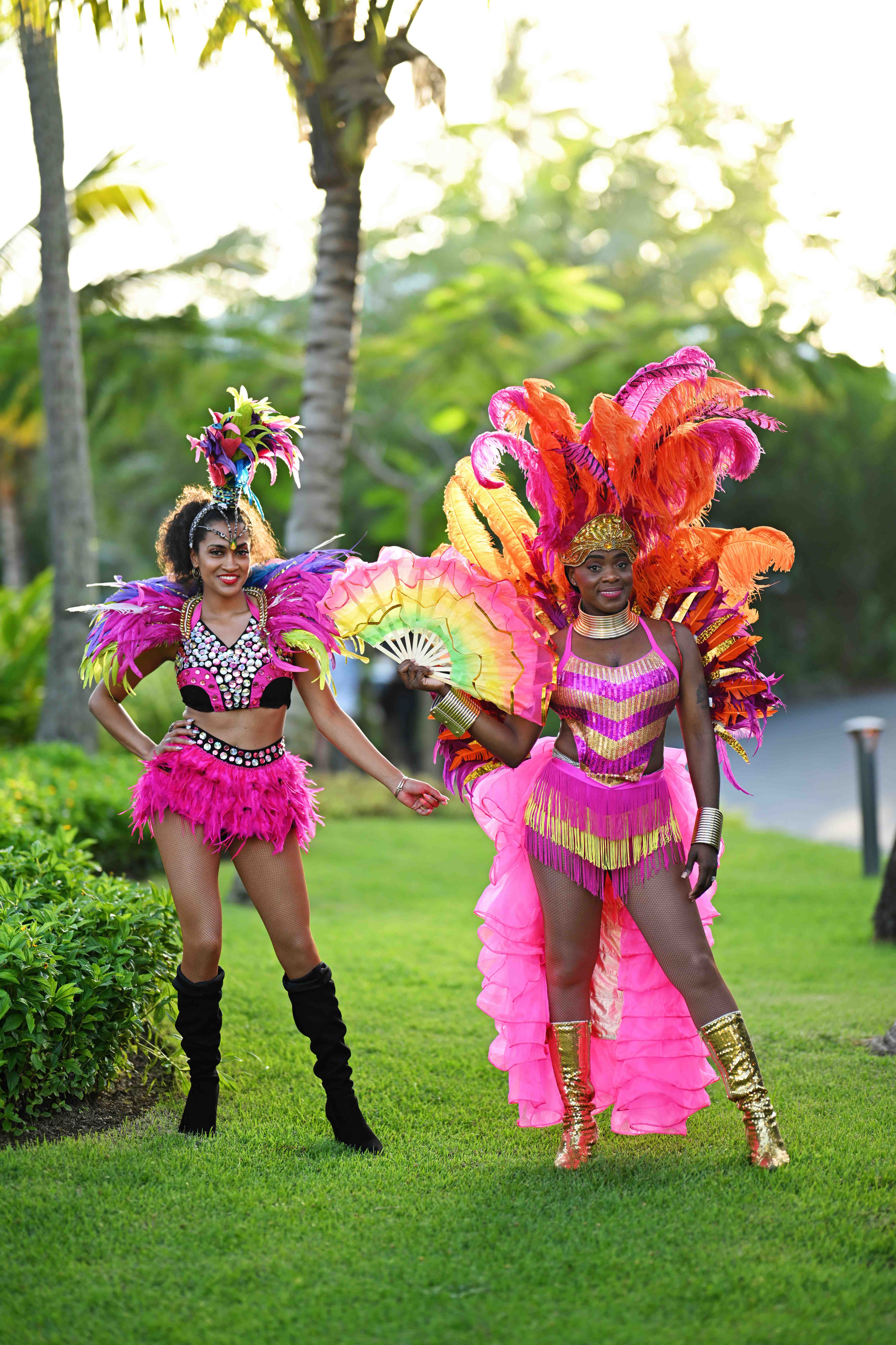 Two Carnival dancers in vibrant pink, orange, and gold feathered costumes at a welcome beach party
